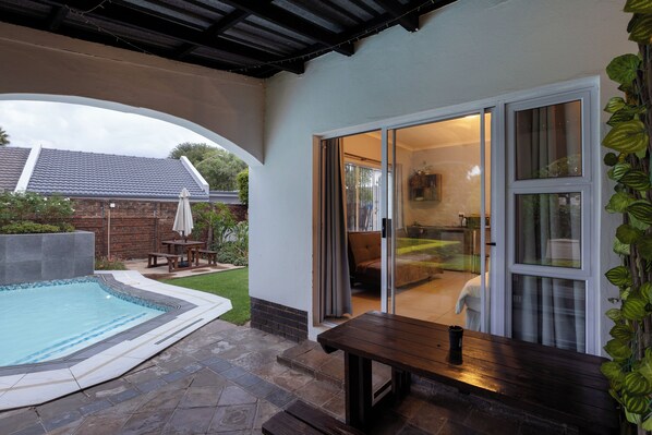 A covered patio with a swimming pool, a wooden bench, and a sliding glass door leading to an interior space with a sofa and a table.