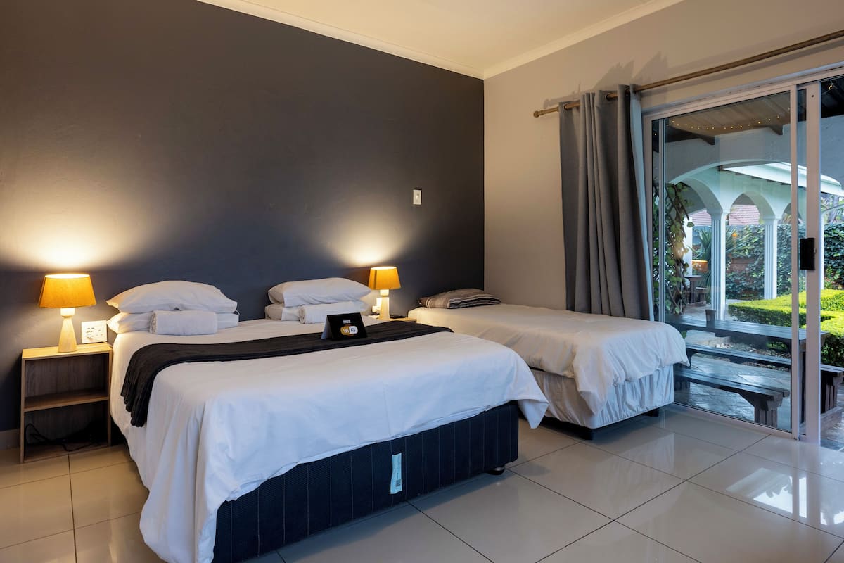 Une chambre d’hôtel avec deux lits, une table de chevet avec une lampe et une vue sur un jardin, visible à travers une porte vitrée.