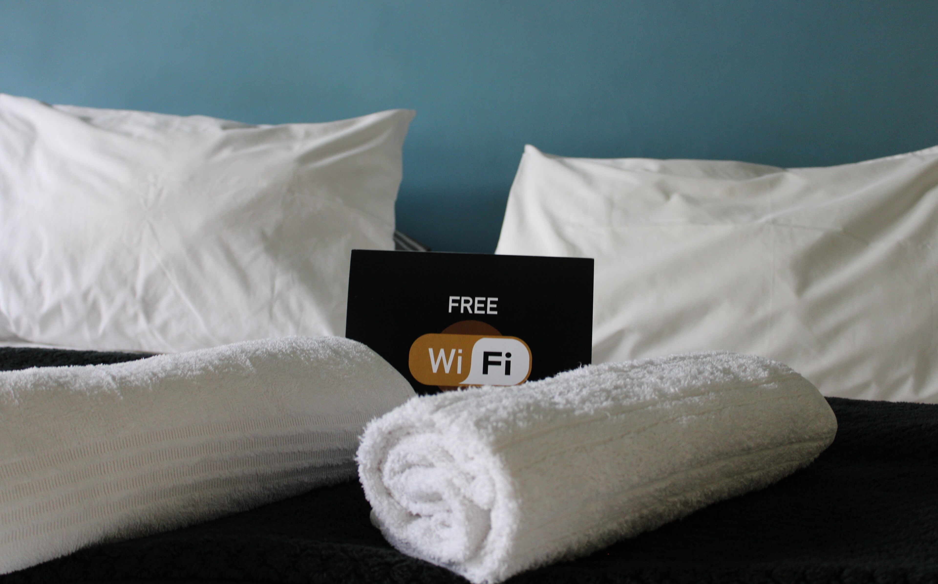 A bed with white pillows and a rolled white towel, a black card with 'FREE WiFi' text, and a blue wall.