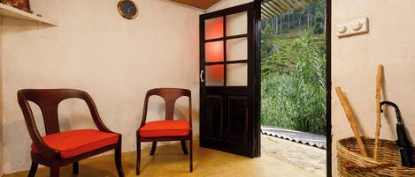 A room with two wooden chairs with red cushions, a black door leading outside, and a wicker basket with sticks and an umbrella.