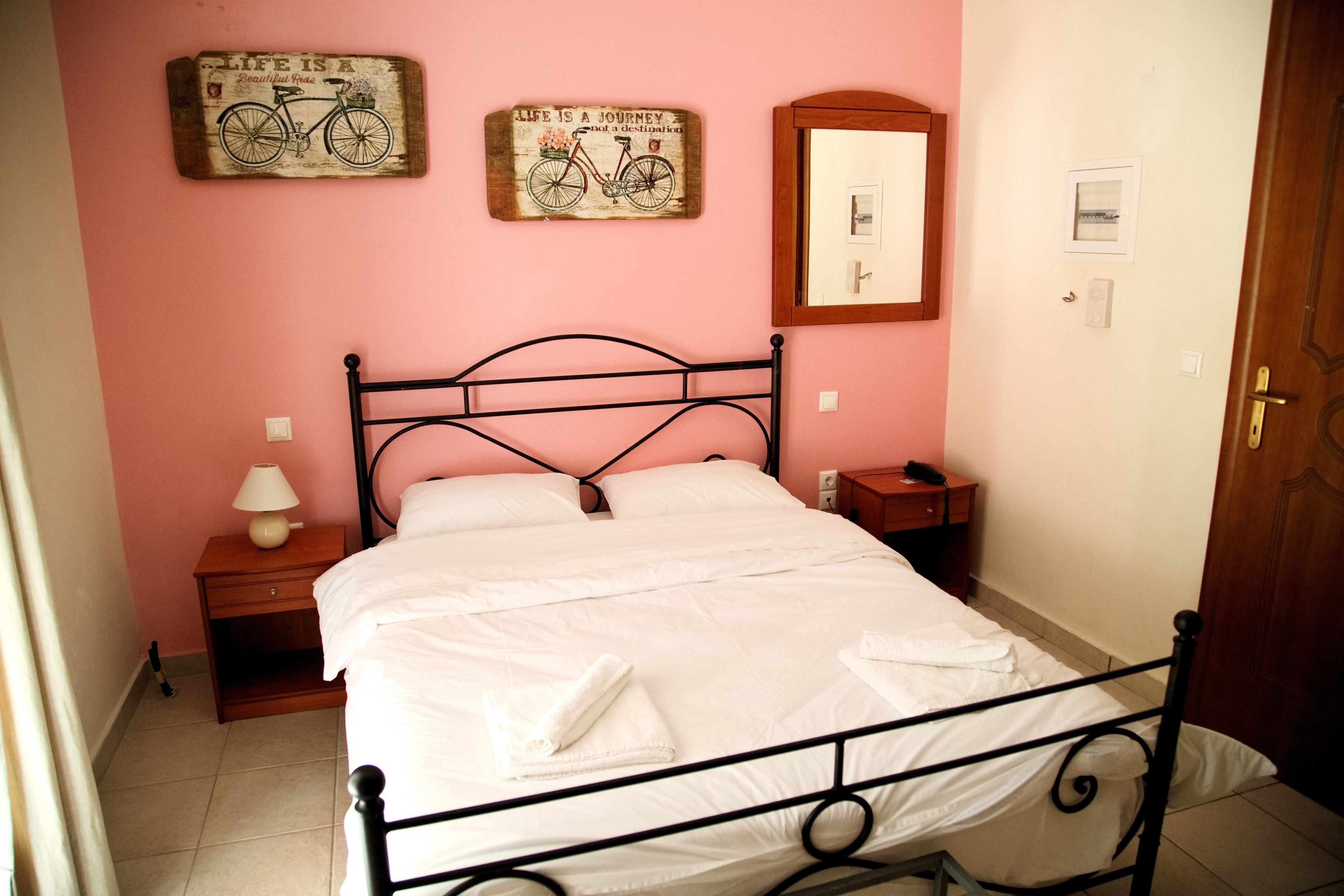 A neatly made bed with white linens and towels, a bedside table with a lamp, and two wall decorations featuring bicycles and a quote.