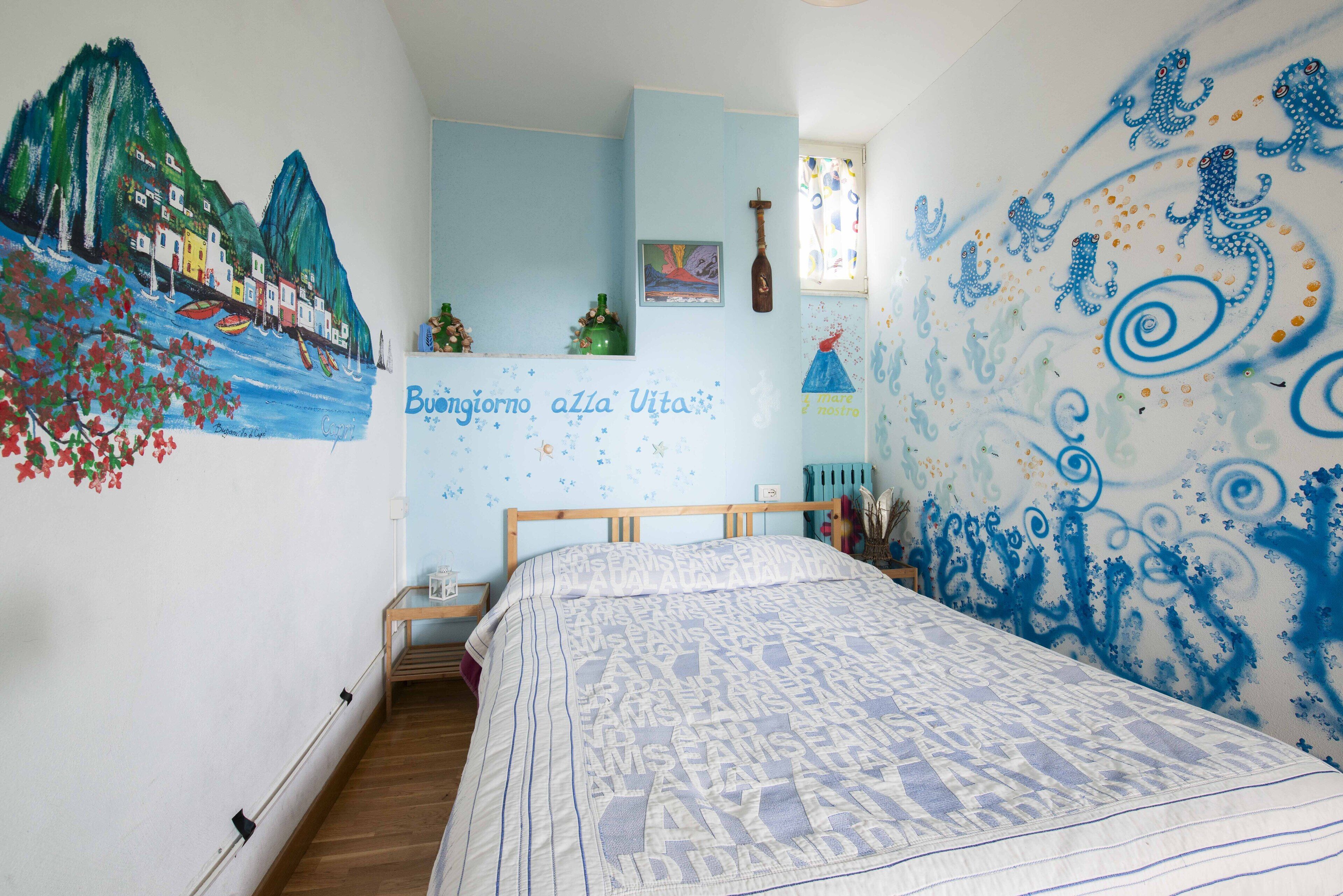Un dormitorio con cama, un mural en la pared y una mesita con una botella y una planta.