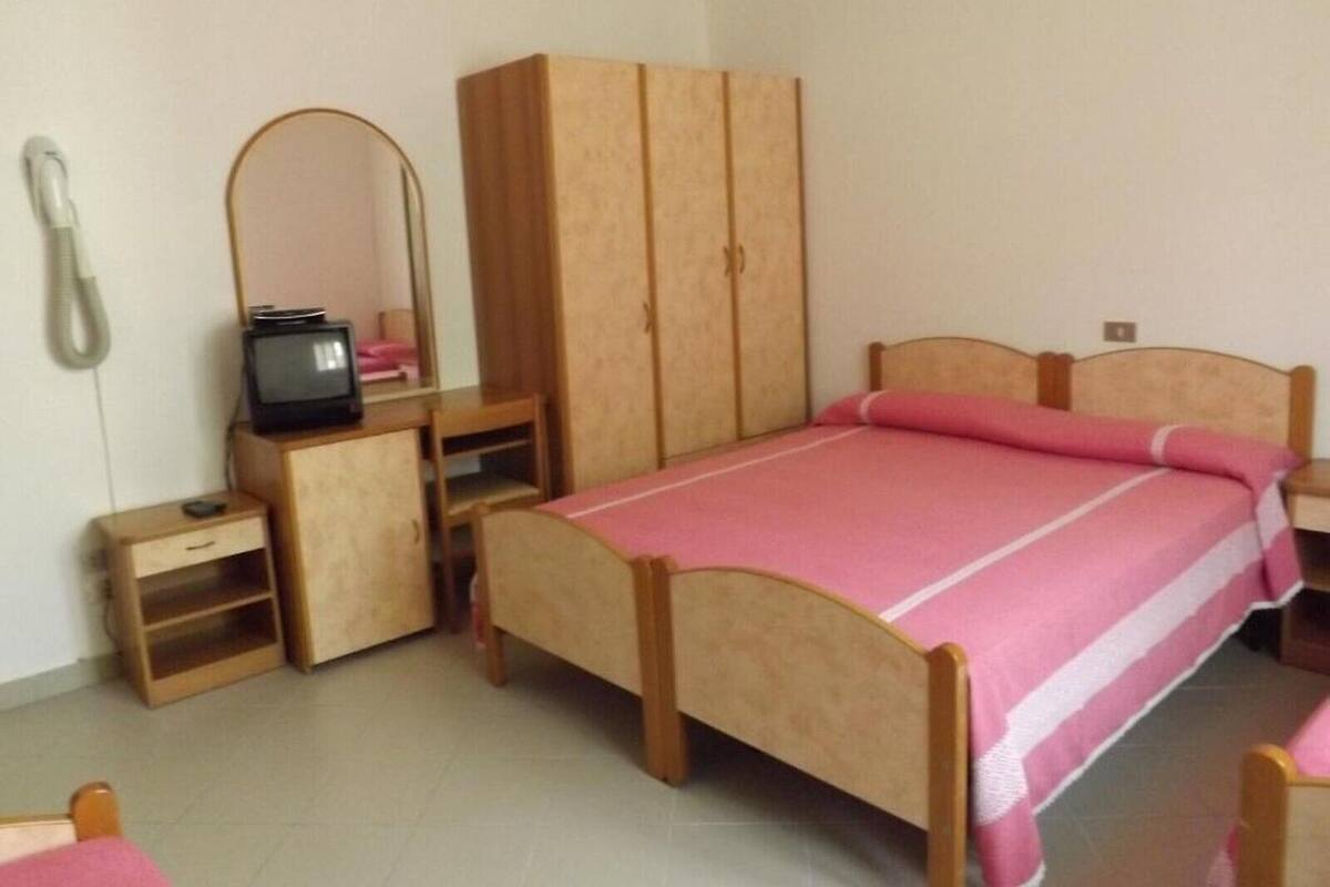A bedroom with a single bed, a wooden wardrobe, a small desk with a chair, a mirror, and a television.