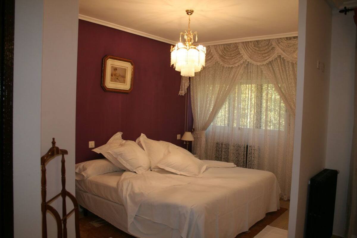 A bedroom with a bed, a chandelier, a window with curtains, and a picture on the wall.
