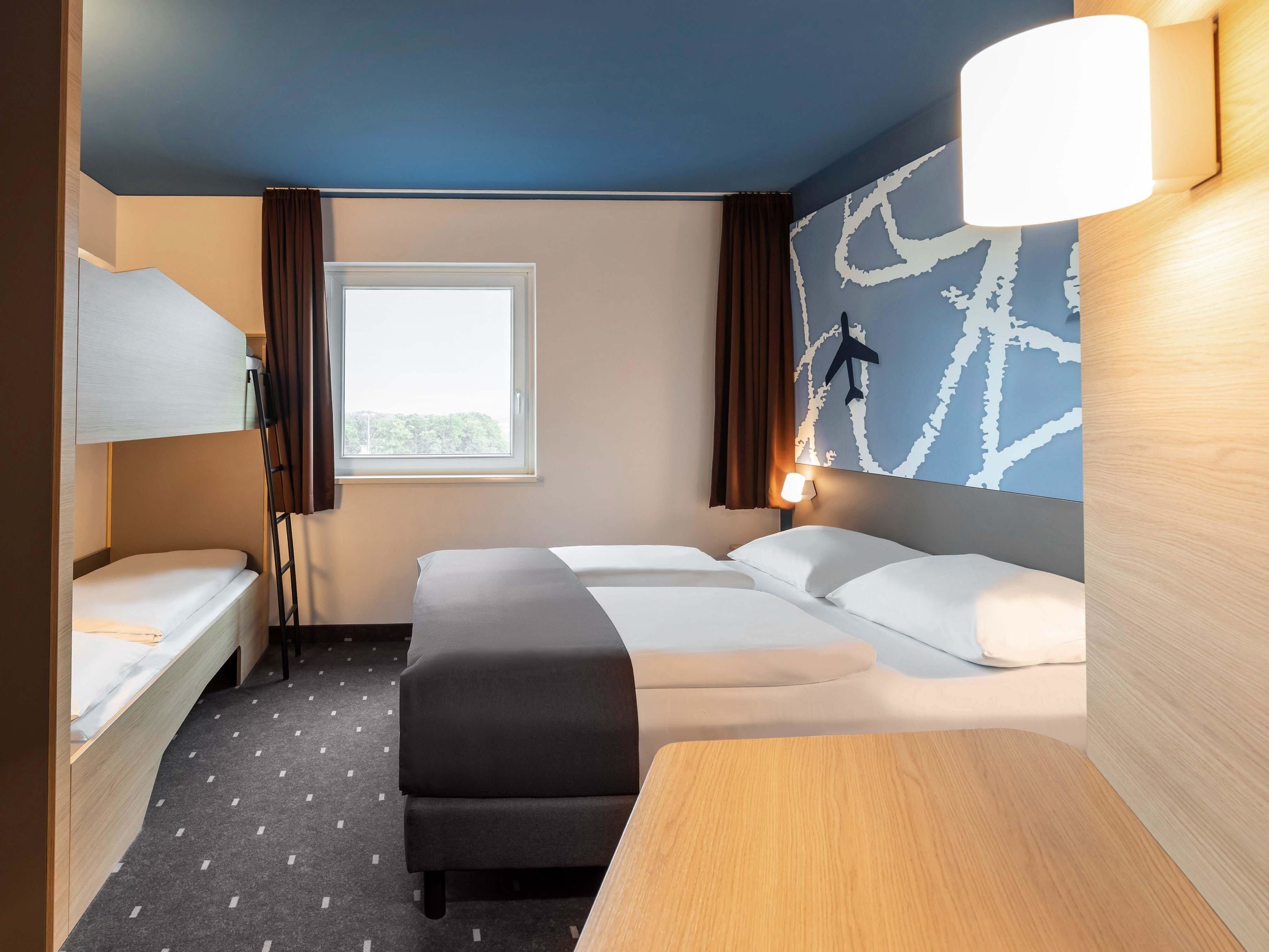 Ein Hotelzimmer mit Etagenbett, einem Einzelbett, einem Schreibtisch und einem großen Fenster.