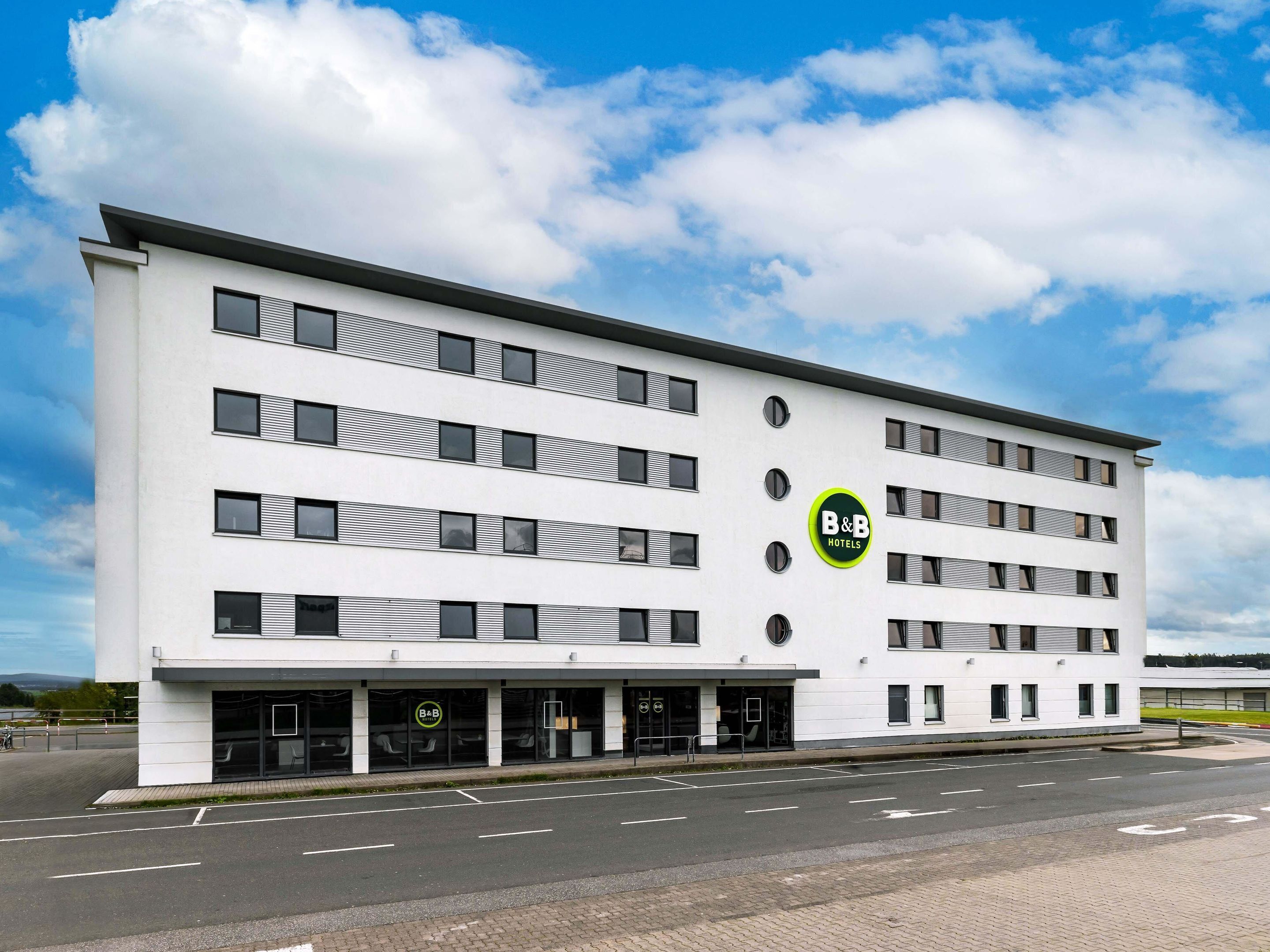 A modern B&B hotel with a white facade, multiple floors, and a parking lot in front.