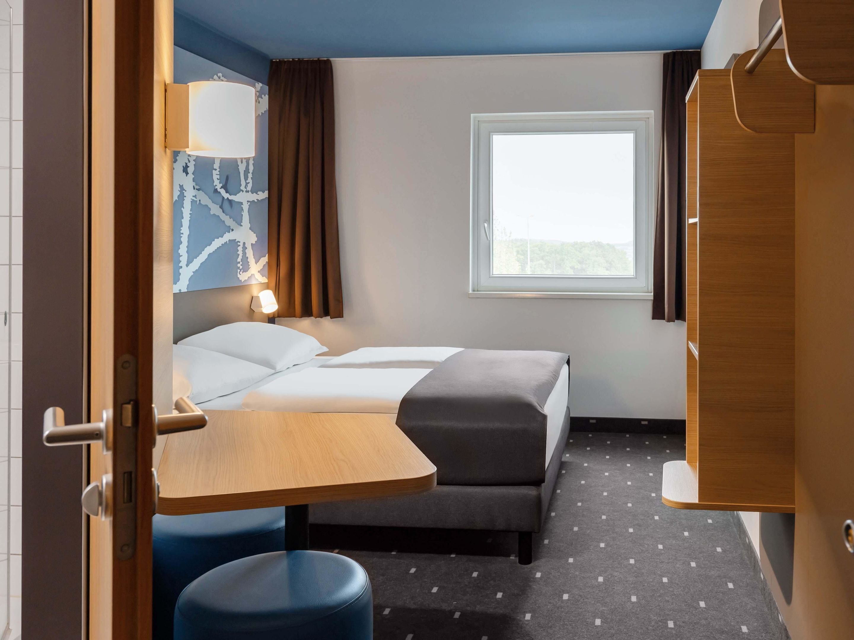 Ein Hotelzimmer mit Bett, Schreibtisch, Stuhl, Fenster und Vorhang.