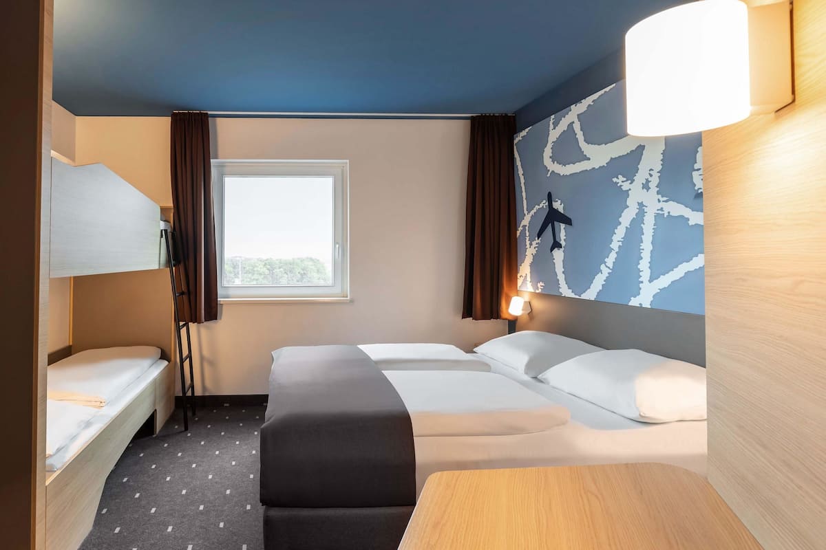 A hotel room with a bunk bed, a single bed, a desk, and a large window.