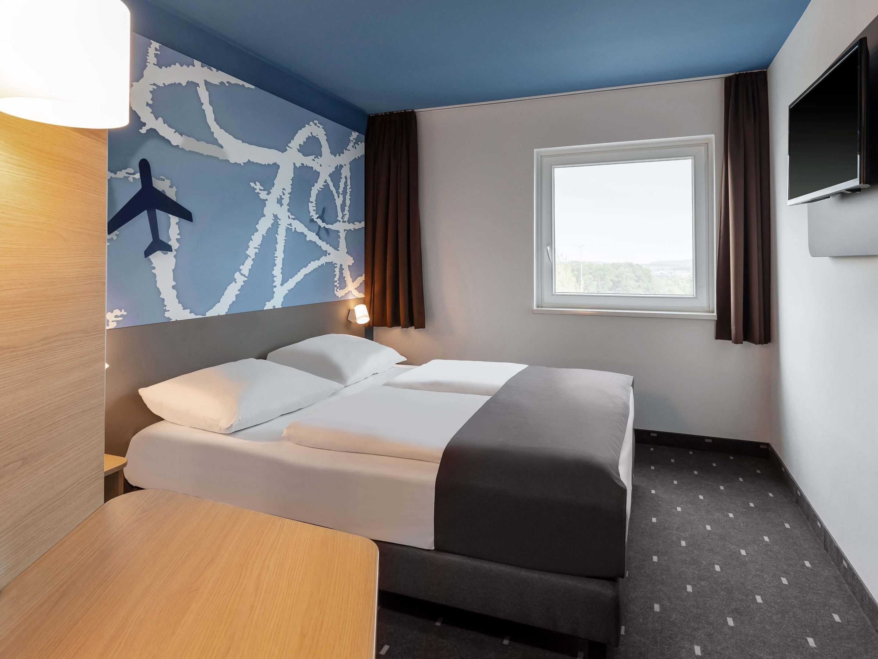 A hotel room with a bed, a desk, a wall-mounted TV, and a window with curtains.