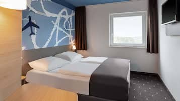 A hotel room with a bed, a desk, a wall-mounted TV, and a window with curtains.