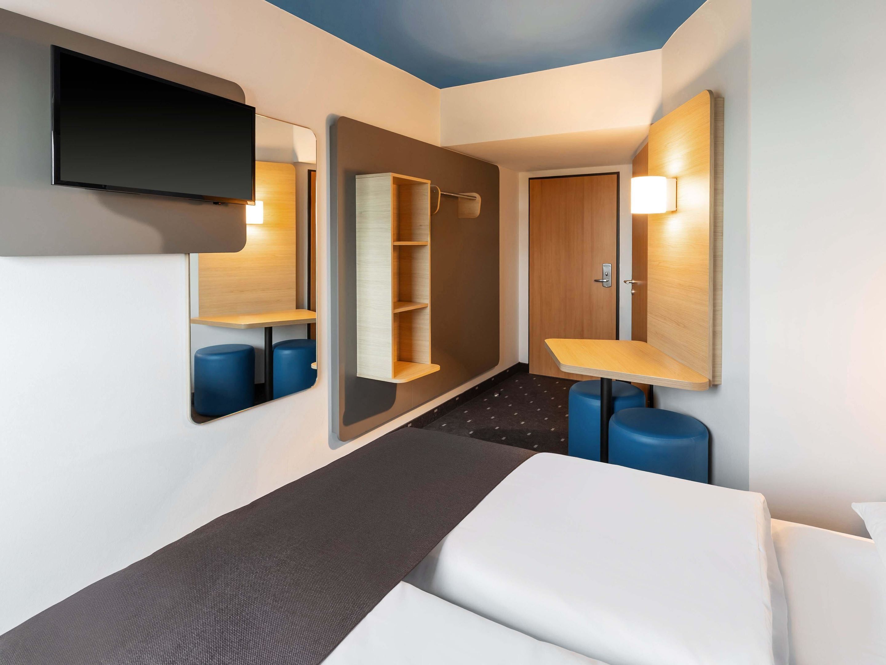 A modern hotel room with a bed, a TV mounted on the wall, a mirror, and a small table with a lamp.