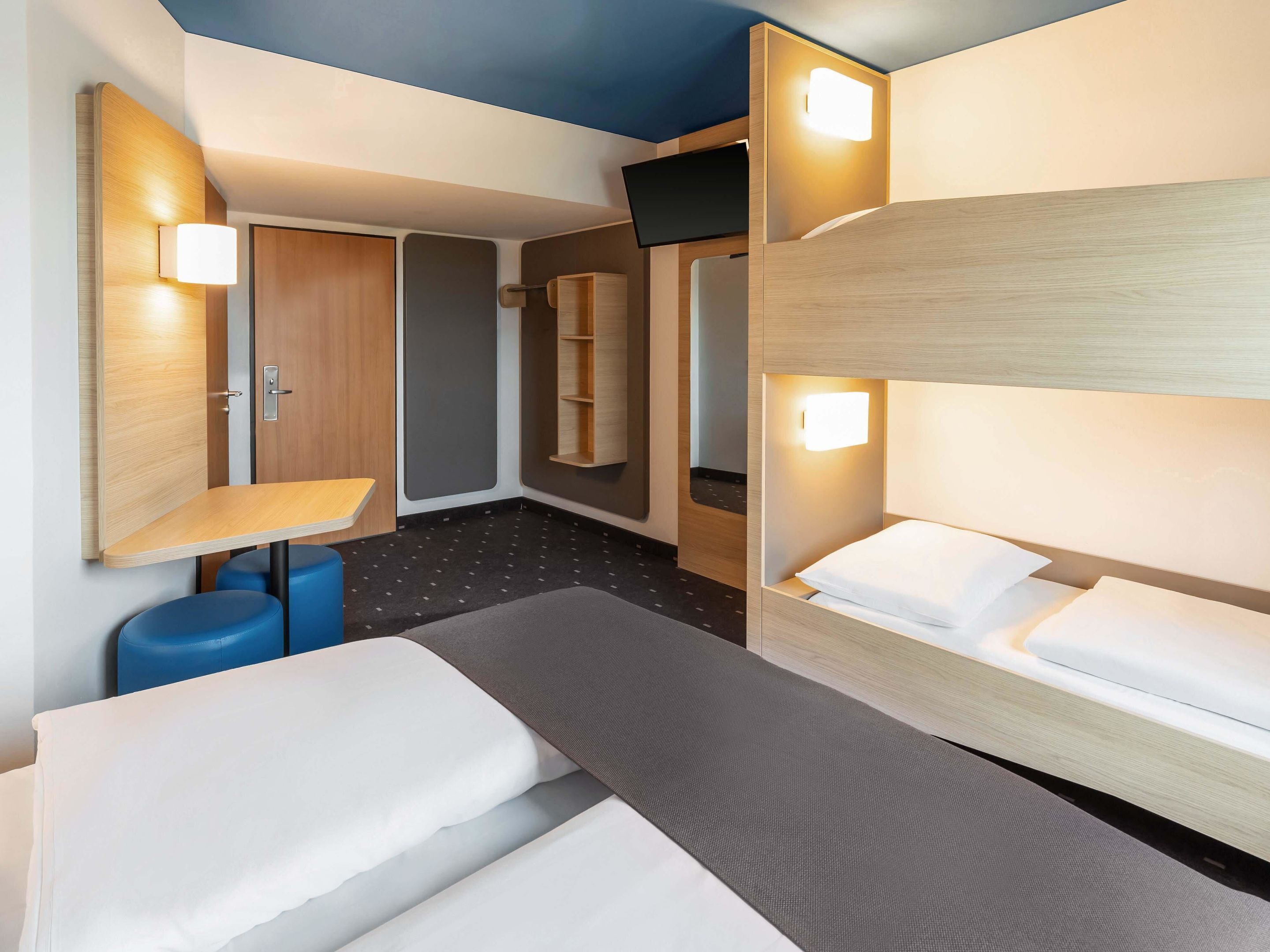 A hotel room with a bunk bed, a desk with a chair, a small table, and a wardrobe.