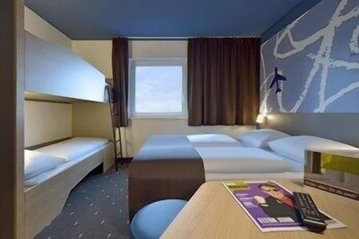 Ett hotellrum med en våningssäng, ett skrivbord med en tidning, en fjärrkontroll och ett fönster med gardiner.