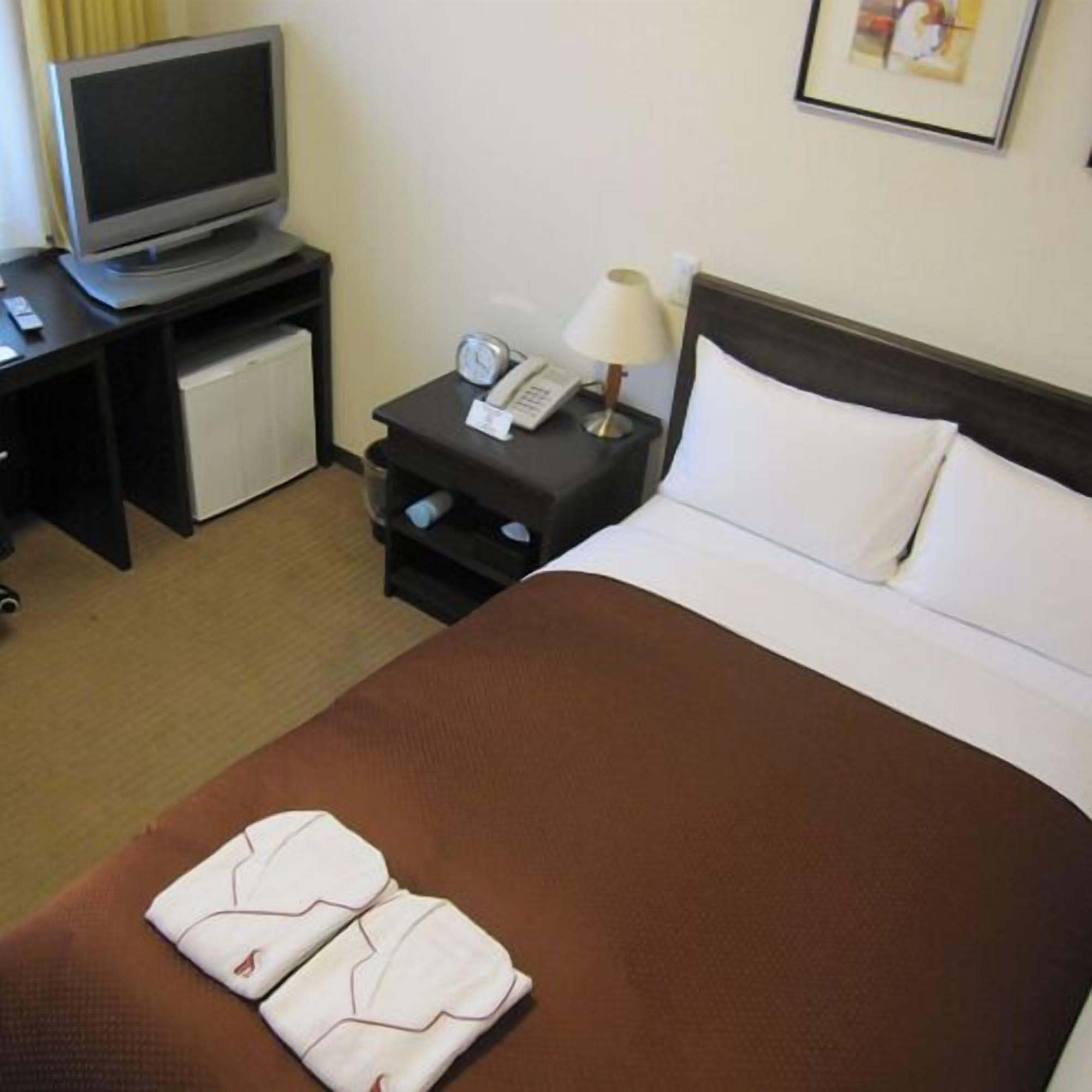 A hotel room with a bed, a TV on a stand, a bedside table with a lamp, and a small fridge.