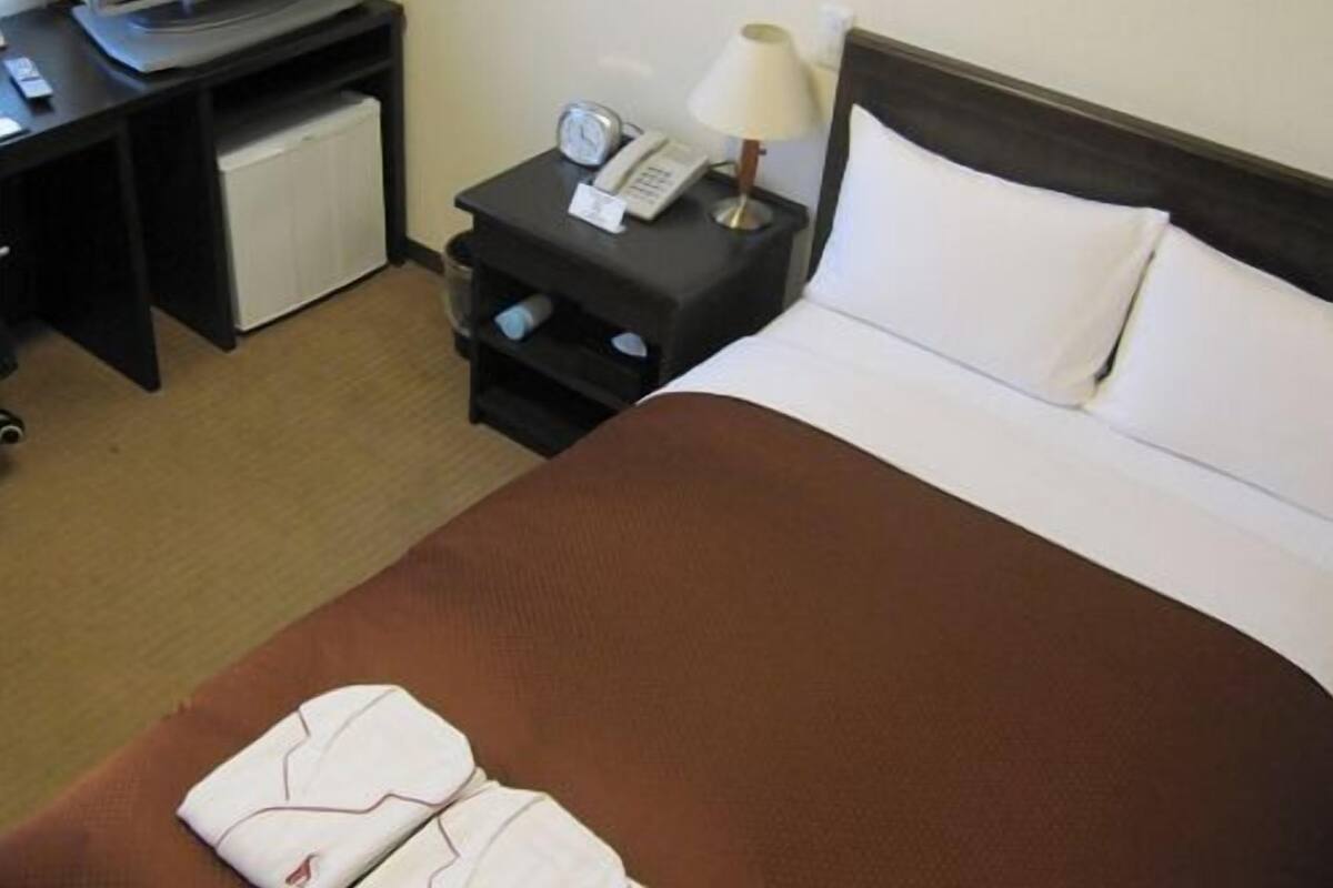 A hotel room with a bed, a TV on a stand, a bedside table with a lamp, and a small fridge.