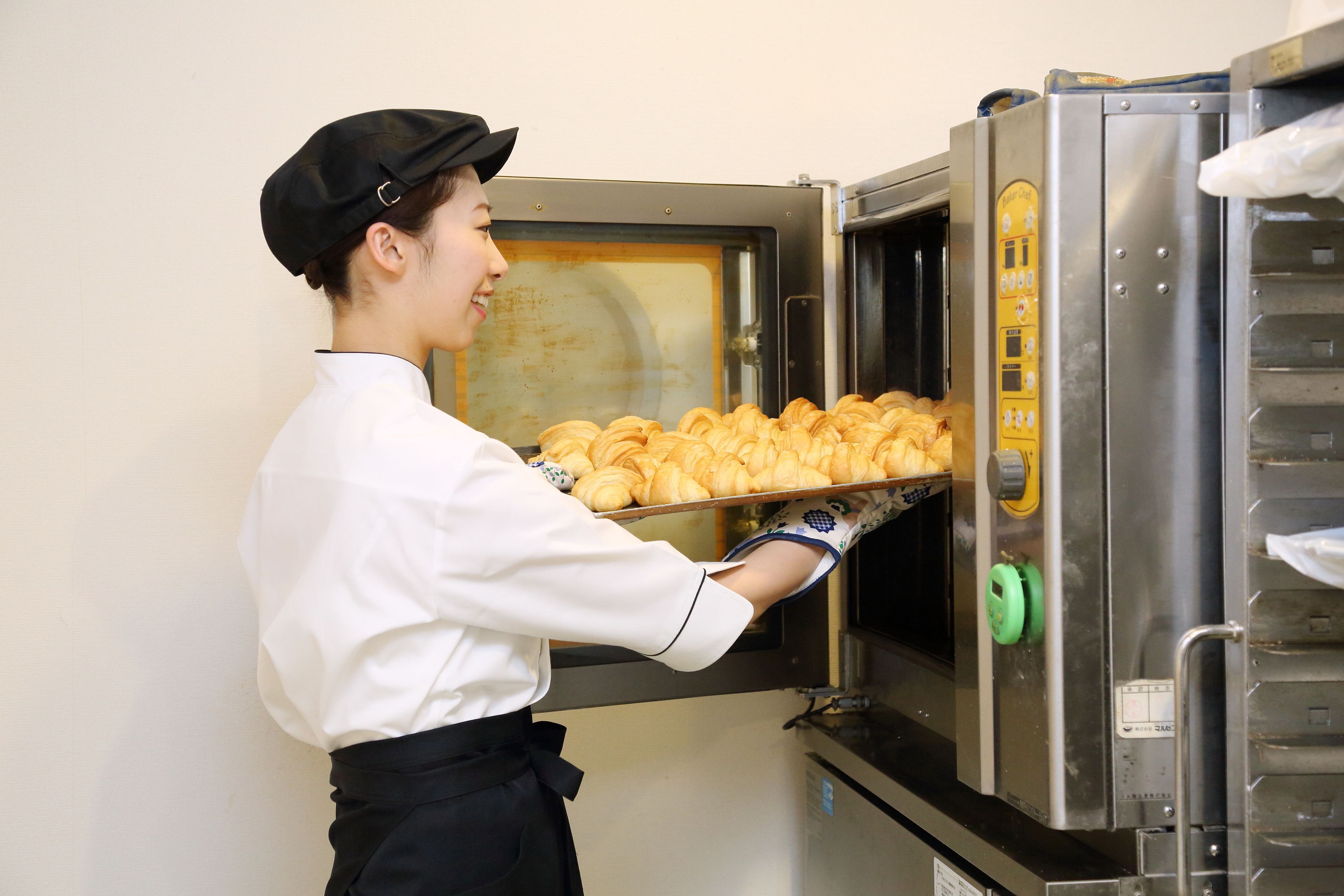 A person in a white shirt and black apron handling a tray of pastries from a commercial oven.