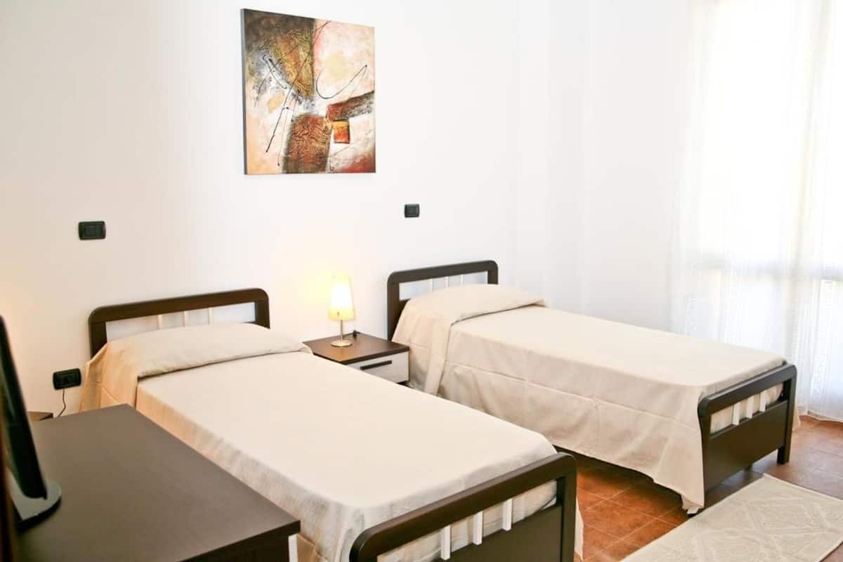 Quarto de hotel com duas camas de solteiro, uma mesa de cabeceira com abajur, um quadro abstrato na parede e uma janela com cortinas.