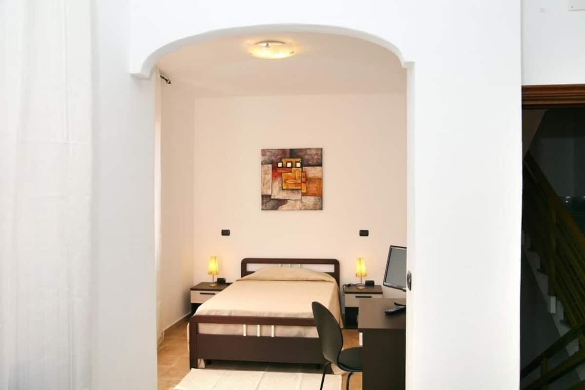 Quarto com cama, luminárias de cabeceira, escrivaninha com computador e um quadro na parede.