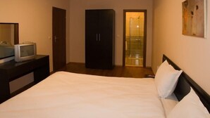 A bedroom with a bed, a TV on a dresser, a wardrobe, and a bathroom visible through a glass door. - Grand Montana (Bansko)