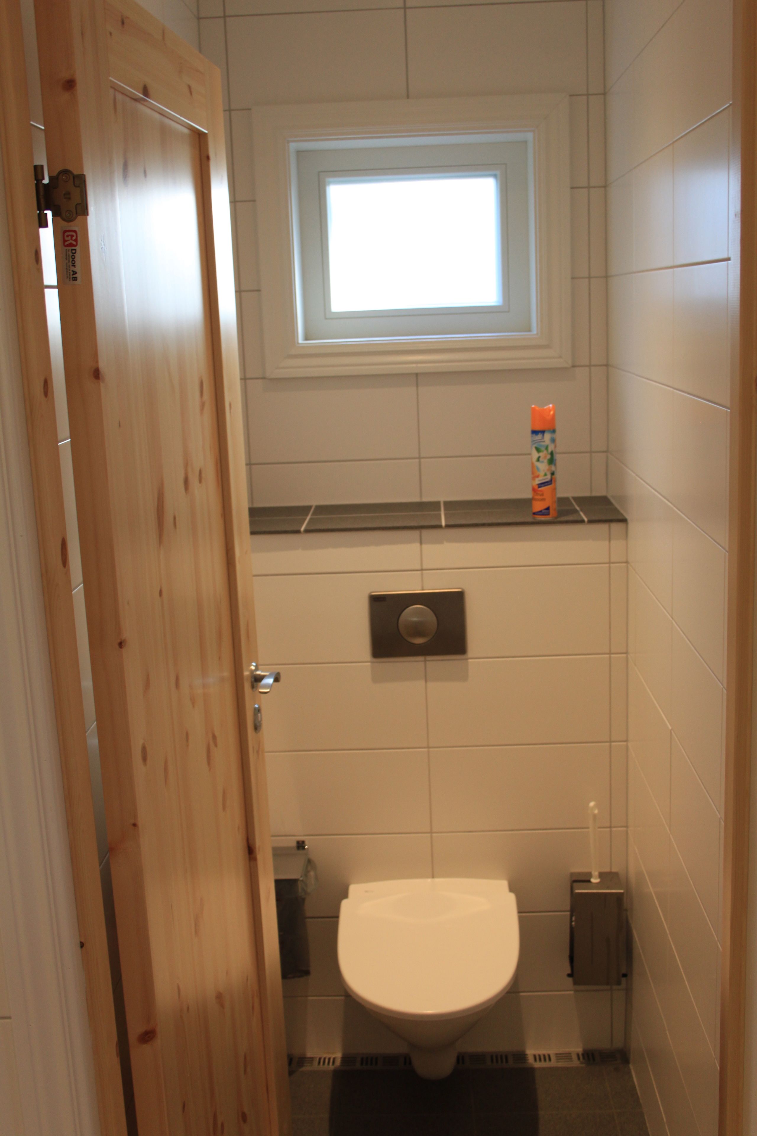 Un baño pequeño y funcional con puerta de madera, ventana sobre el inodoro y repisa con una botella.