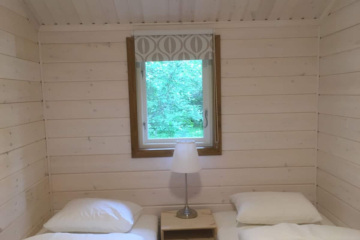 A small, cozy room with two beds, a nightstand with a lamp, and a window with a view of greenery.