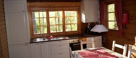 Eine Küchenzeile im Chalet-Stil mit Holzwänden, einem Fenster mit vielen kleinen Scheiben, weißen Schränken, einer Spüle, einem Herd und einem Esstisch mit rotem kariertem Tischläufer.
