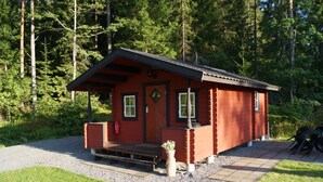 A small, red cabin with a wooden porch and a fire extinguisher, surrounded by a forest. - Långsjön Stugor & Camping (Ankarsrum)