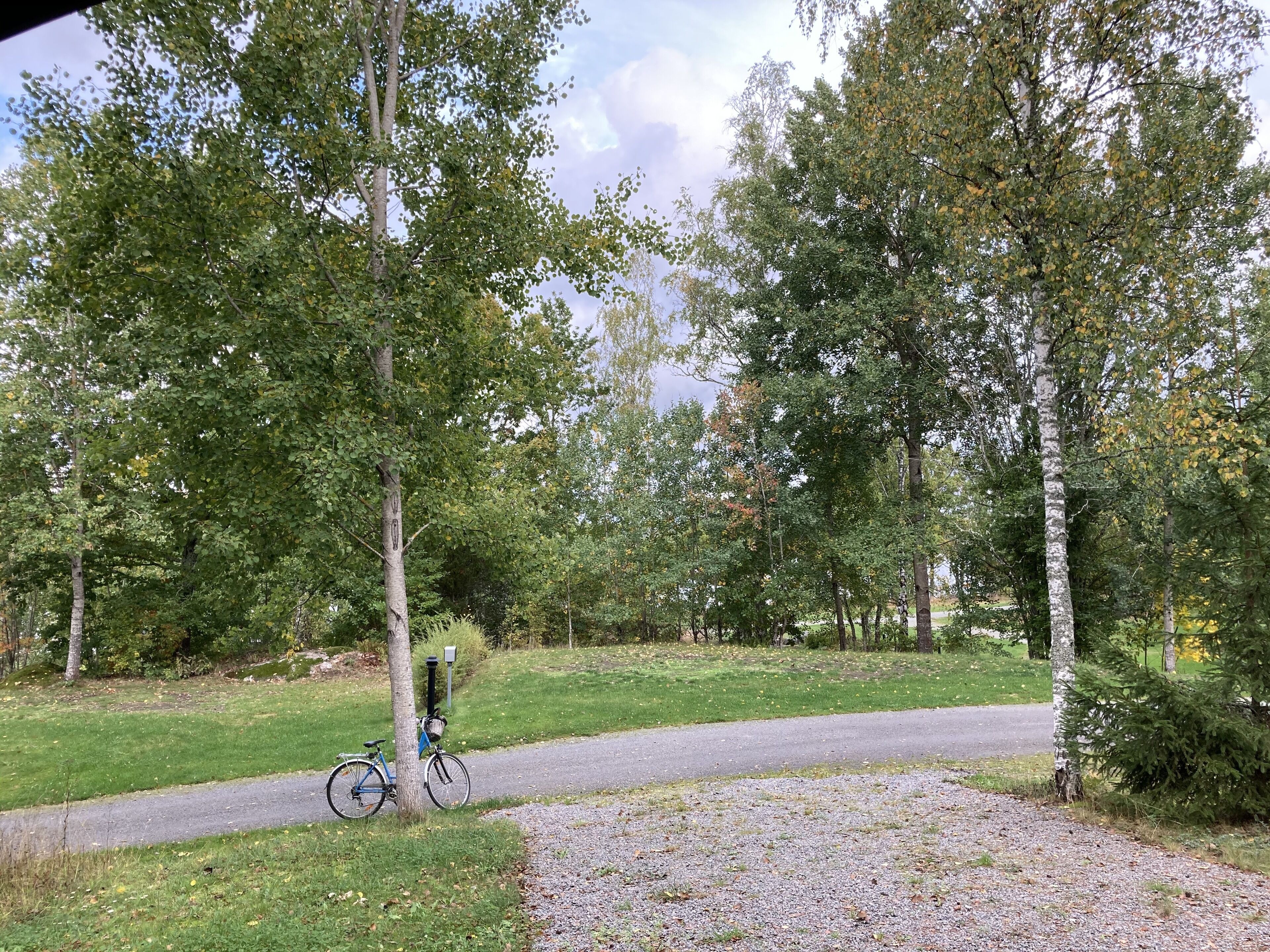 Un chemin de gravier traverse un parc arboré, et un vélo est garé sur le bord.