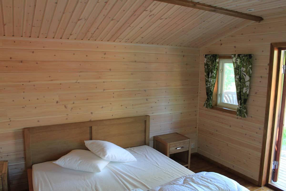 A wooden cabin room with a bed, a nightstand, and a window with curtains.