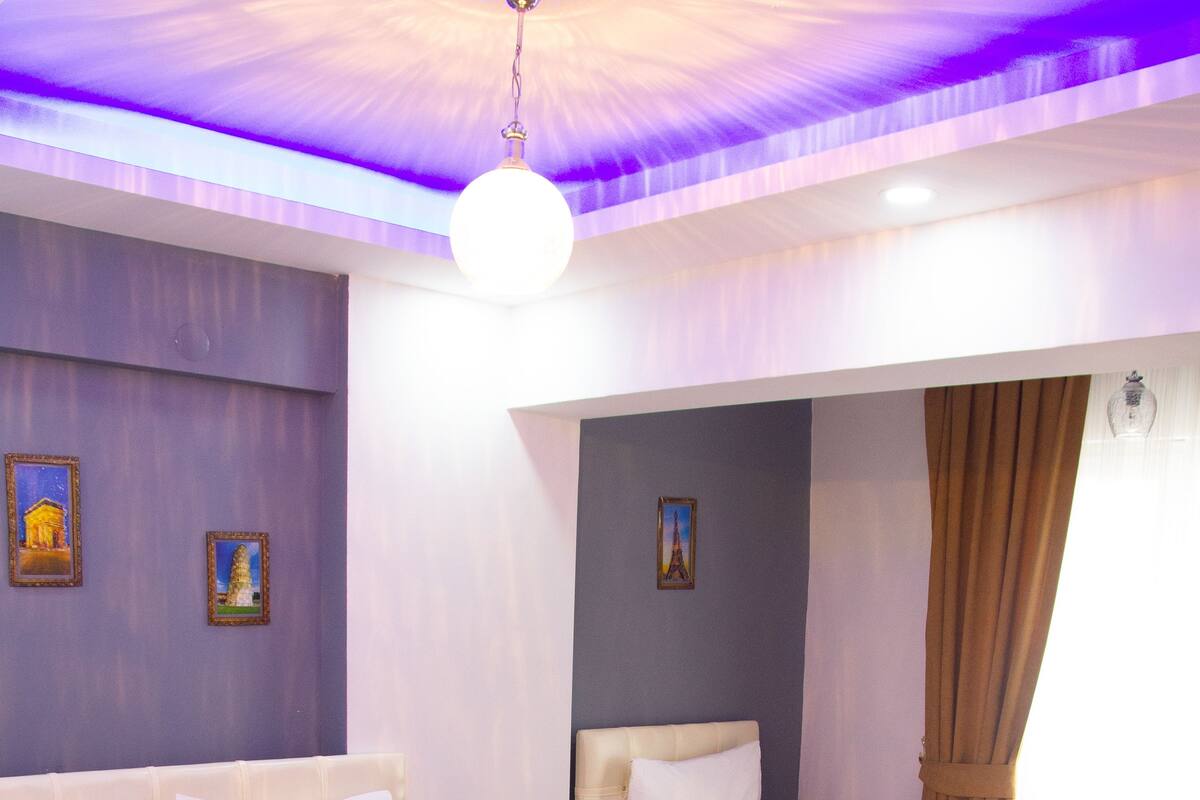 Une chambre d’hôtel avec deux lits, chacun recouvert d’une couverture marron et agrémenté d’oreillers blancs. Les murs sont ornés de tableaux encadrés, il y a un luminaire au plafond et une fenêtre avec des rideaux.