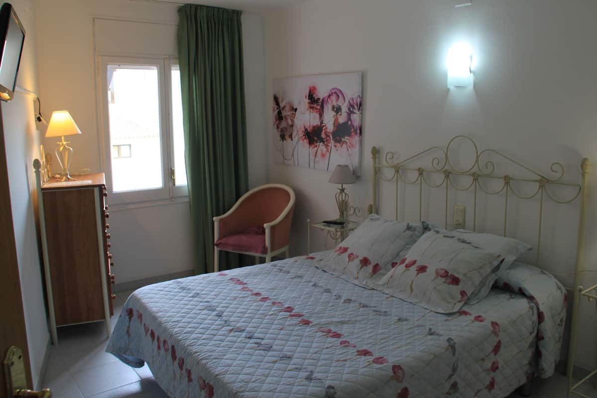 A bedroom with a bed, a chair, a bedside table, a lamp, and a painting on the wall.