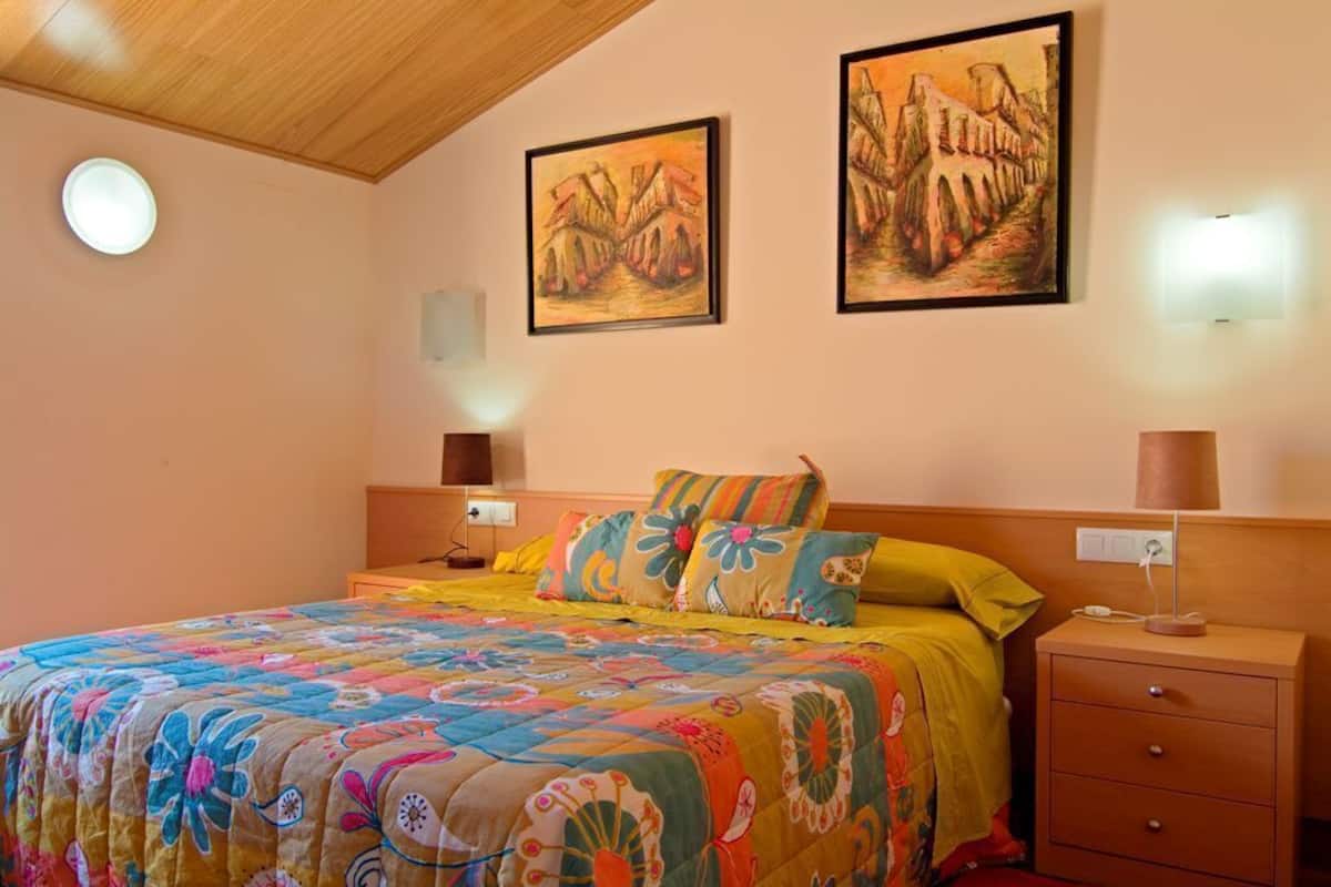 A bedroom with a bed, two wall-mounted paintings, a bedside table with a lamp, and a nightstand.