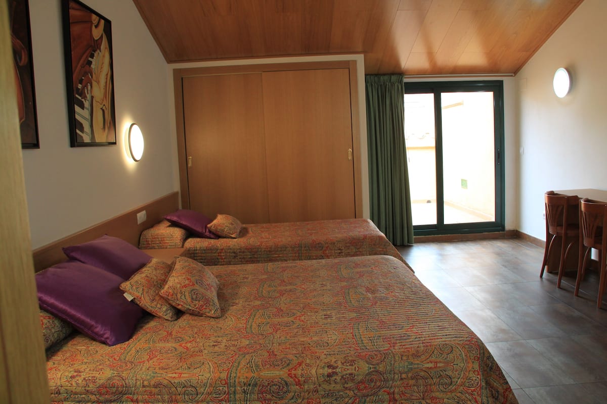 A room with two beds, a wooden ceiling, a sliding door, a dining area with chairs, and a view through the door.