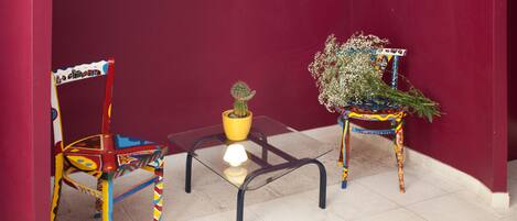 A room with a red wall, a glass table with a yellow pot and a small lamp, and two colorful chairs.