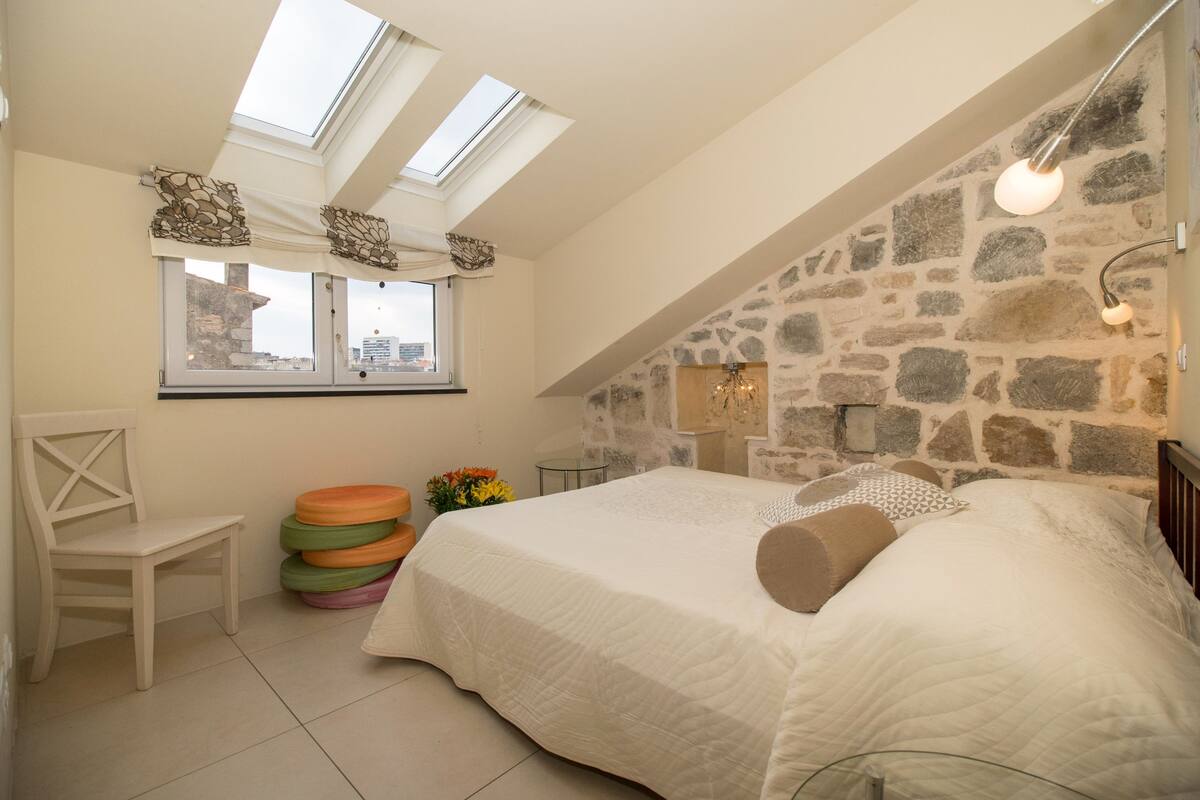 A bedroom with a sloped ceiling, stone wall, a bed with white bedding, a chair, and a window with curtains.