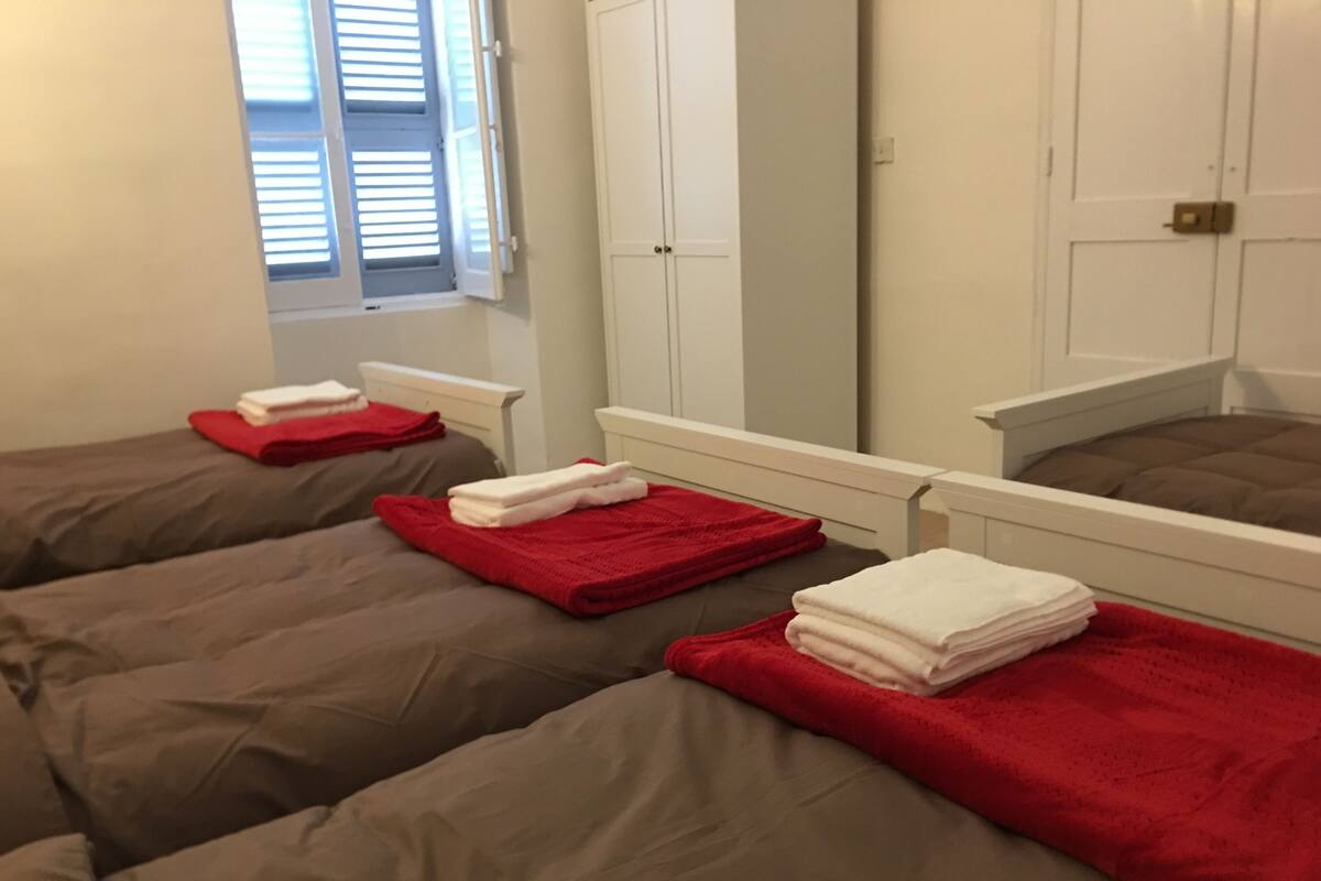 Une chambre d’hôtel avec deux lits, chacun agrémenté d’une serviette rouge et d’une serviette blanche pliée et posée sur le dessus. On y trouve une fenêtre avec des stores, une armoire blanche et un miroir au mur.