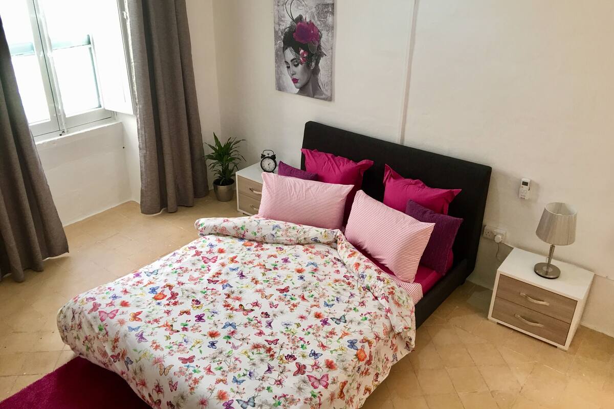 Une chambre à coucher avec une couvre-lit à motifs floraux, une tête de lit noire et des coussins roses. On y trouve une table de chevet avec une lampe et une horloge.
