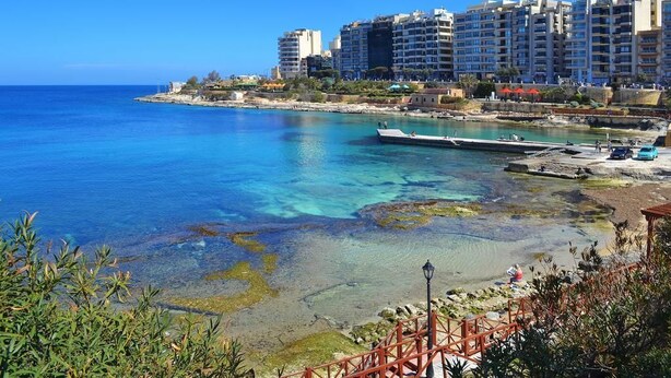 Una zona costera con aguas azules cristalinas, un muelle y una hilera de edificios de varios pisos al fondo.