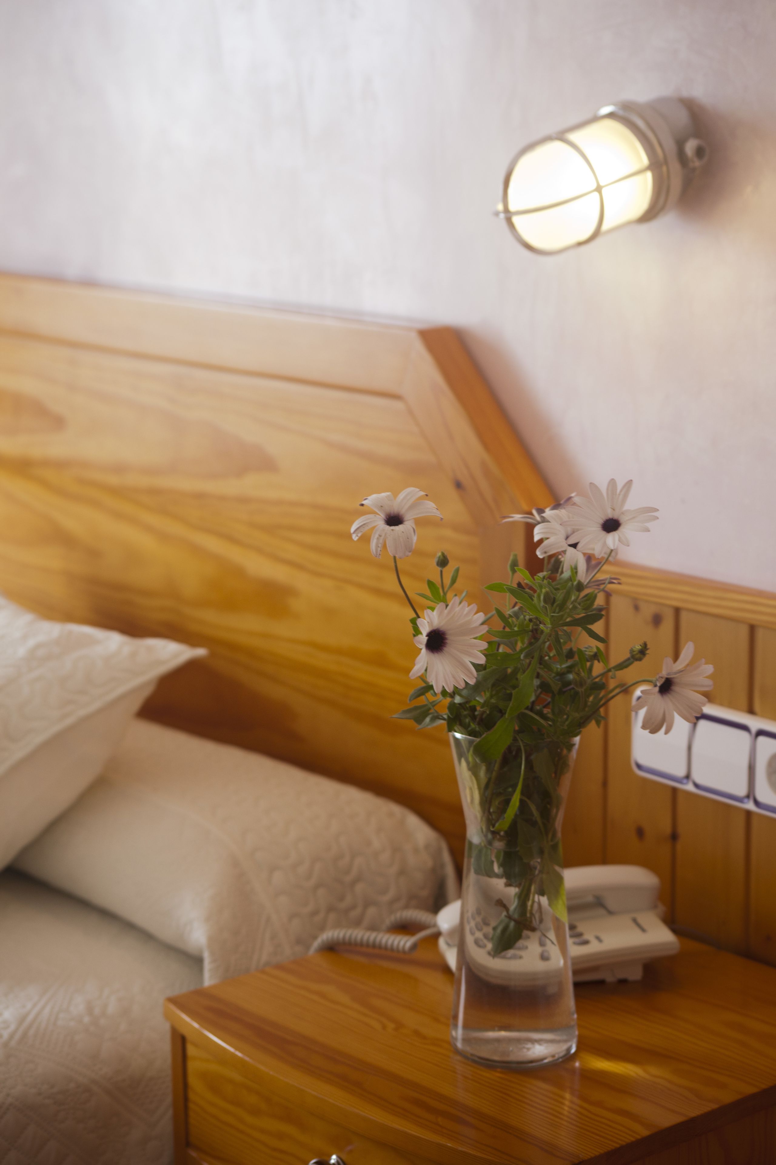 Ein Nachttisch aus Holz mit einer Vase voller weißer Blumen, einem Telefon und einer Wandleuchte in einem Hotelzimmer.