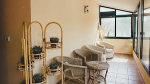 A sunlit room with wicker furniture, a rattan plant stand, and a view through large windows. - B&B Villa Grazia (Alghero)