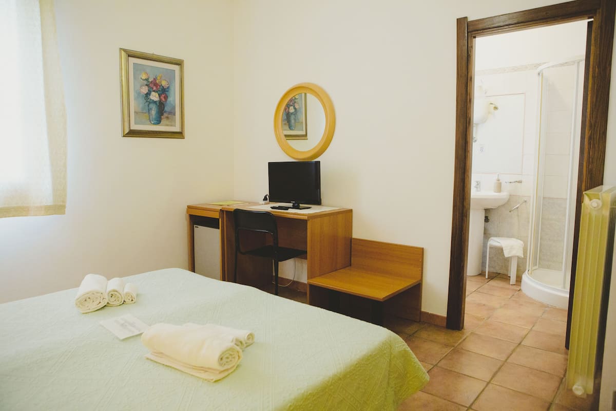 Habitación de hotel con cama, escritorio con computadora, silla, taburete pequeño, espejo y baño con ducha.