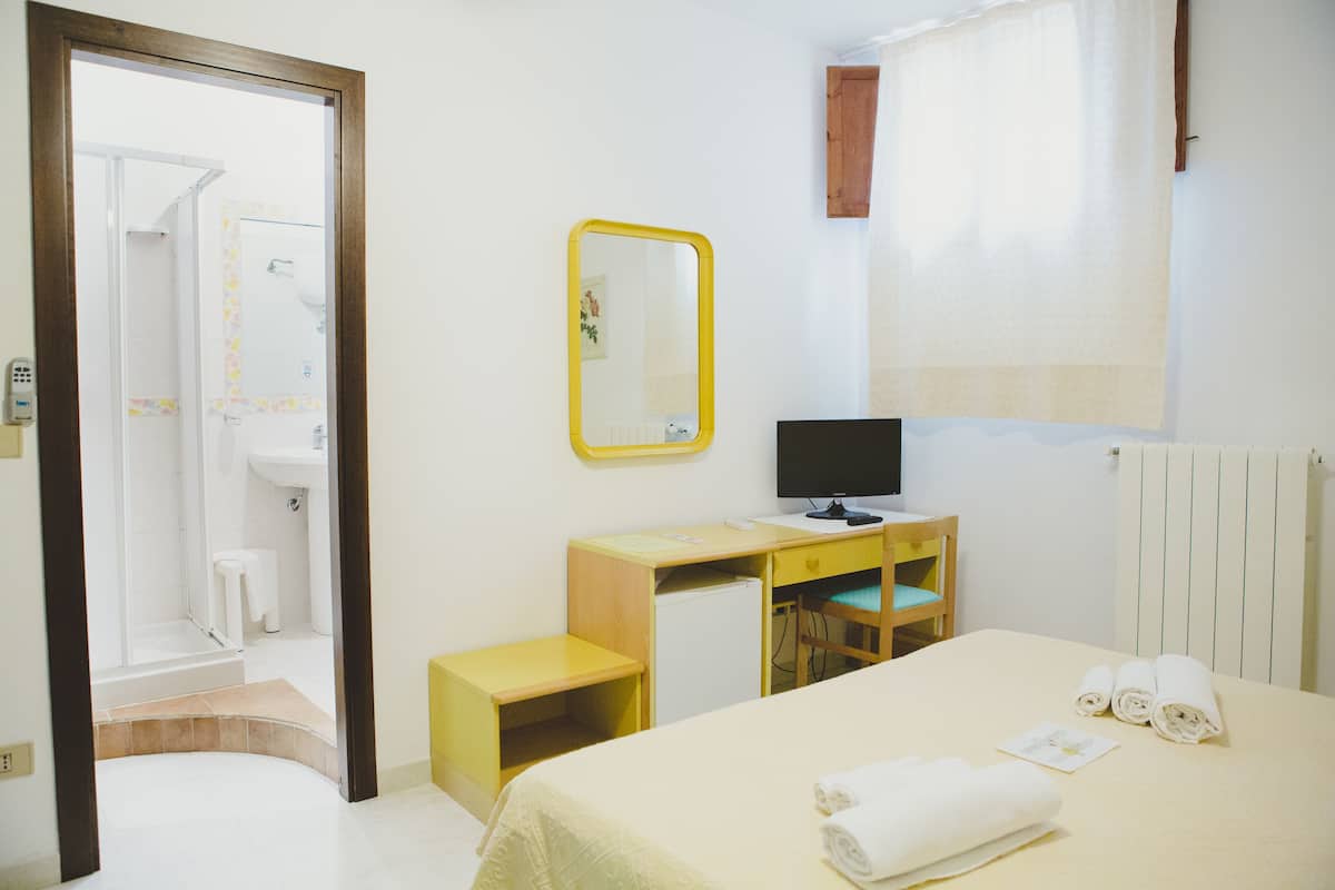 Habitación de hotel con cama, escritorio, silla, televisor, espejo y baño visible a través de una puerta abierta.