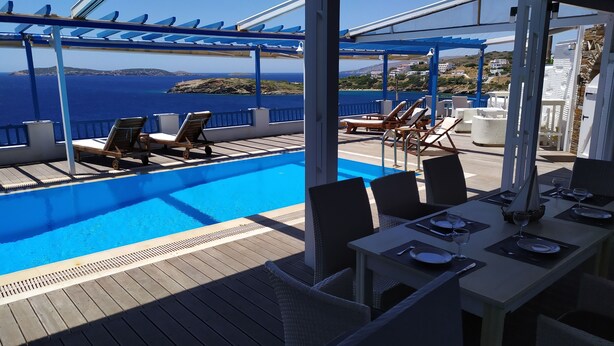 A poolside area with lounge chairs and a table set for dining, overlooking a clear blue sea and a hilly landscape. - Blue Waves View (Andros)