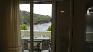 A view from inside a room to a balcony with a table and chairs, overlooking a rocky coastline. - Blue Waves View (Andros)