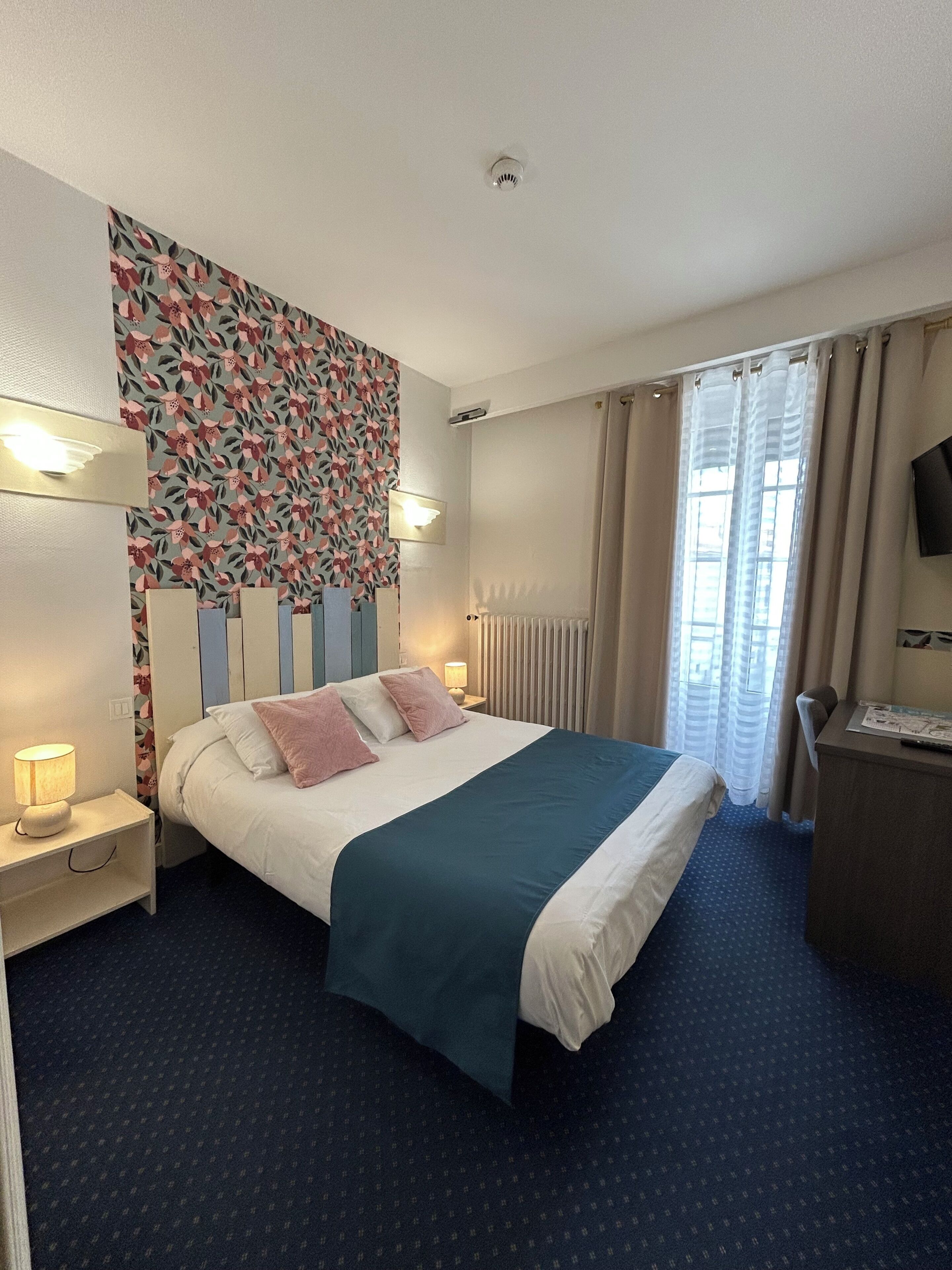 A hotel room with a bed, bedside lamps, a desk, a television, and a window with curtains.