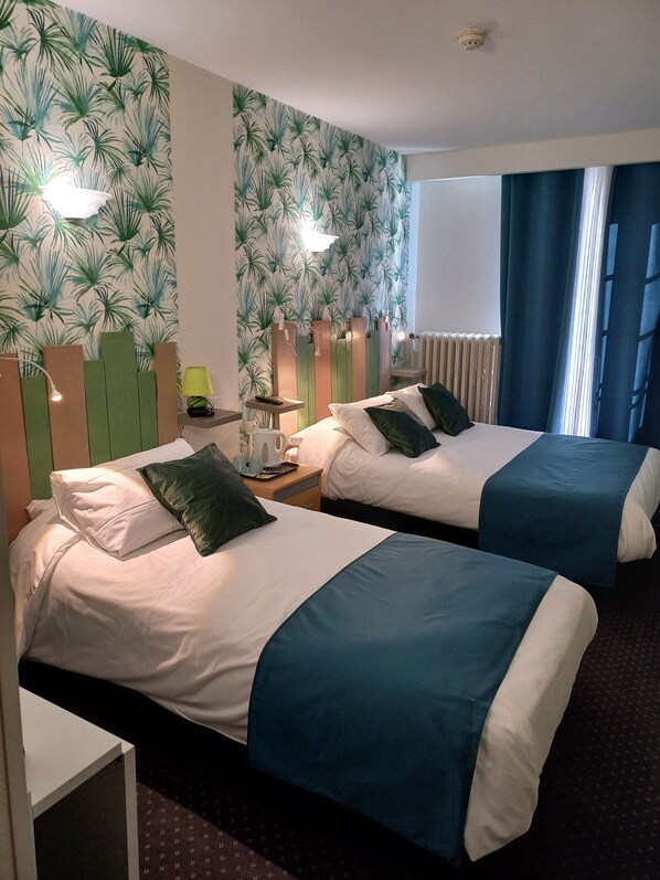 A hotel room with two beds, a small table, a lamp, and a radiator.