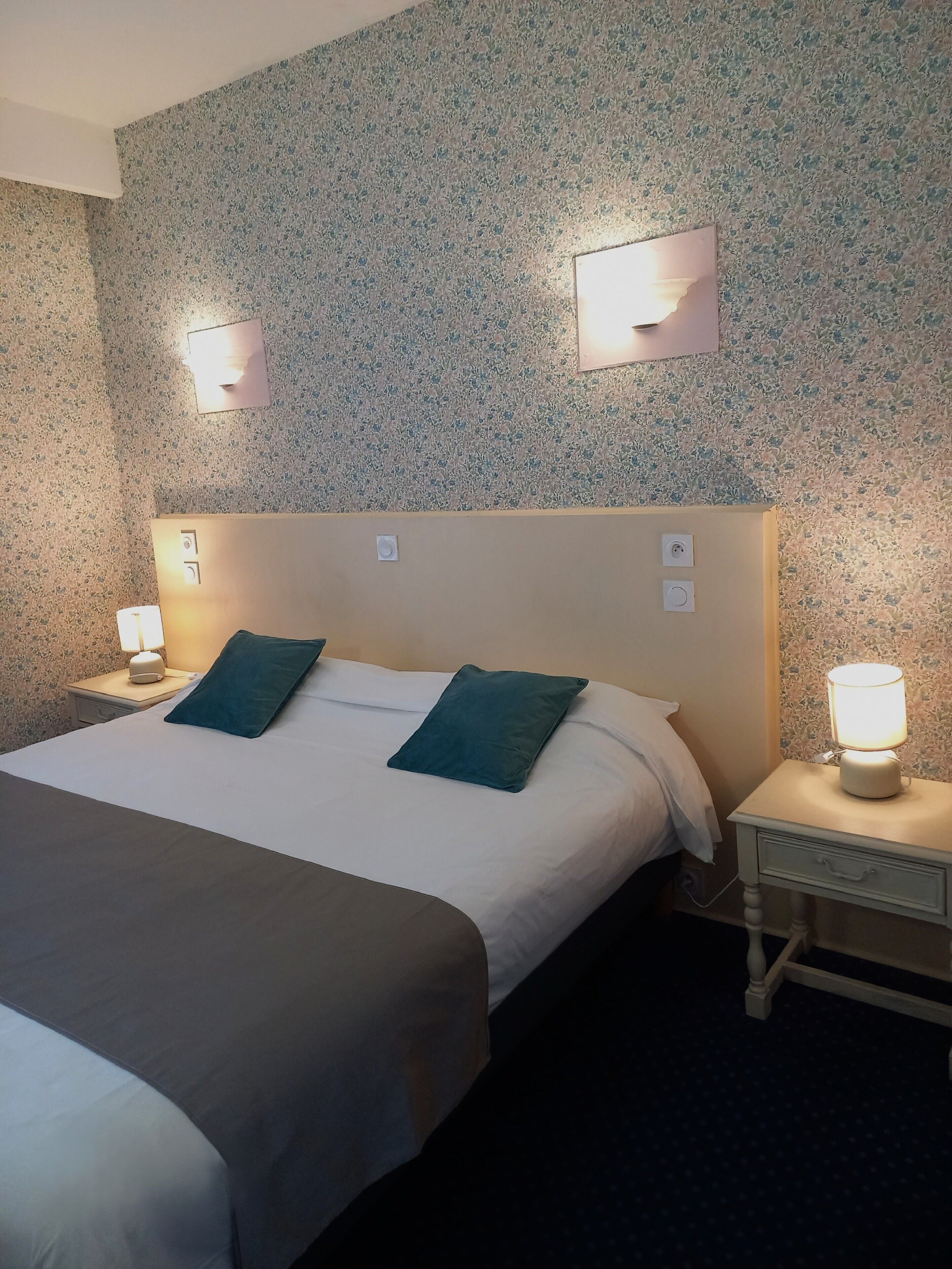 A hotel room with a bed, two pillows, a nightstand with a lamp, and a wall-mounted light fixture.