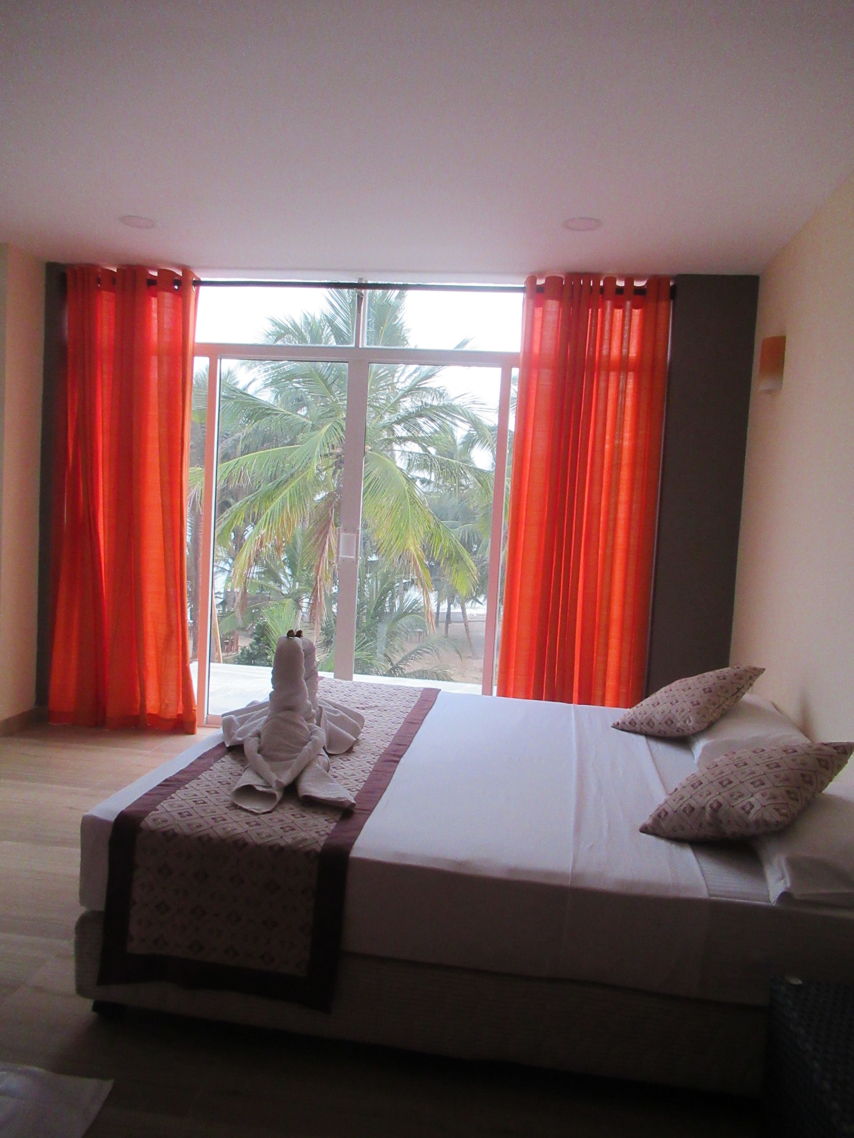 Un dormitorio con una cama grande, dos almohadas y una toalla sobre la cama. Hay una ventana con cortinas y vistas a palmeras desde el exterior.
