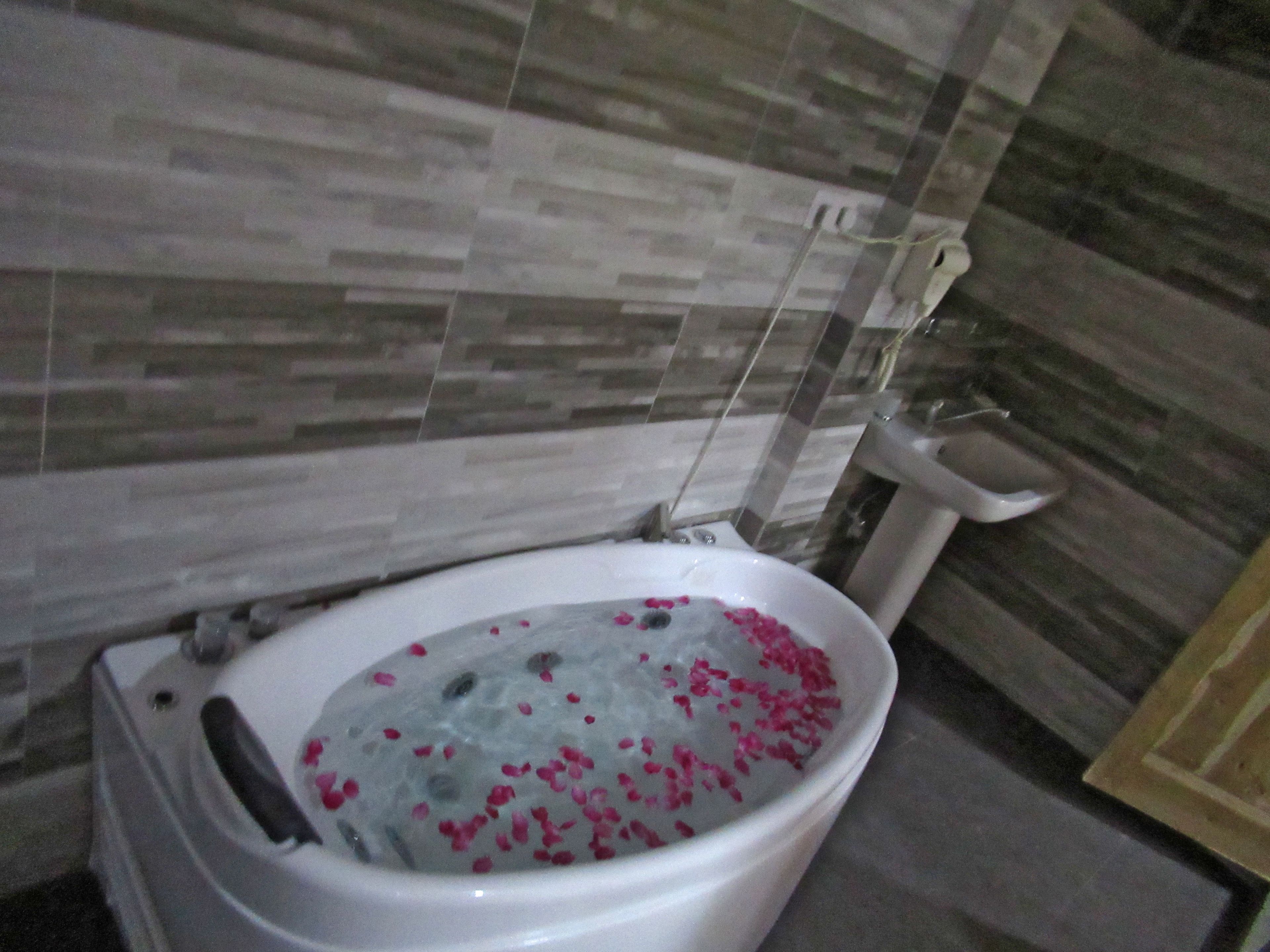 A bathroom with a bathtub filled with water and rose petals, a toilet, and tiled walls.