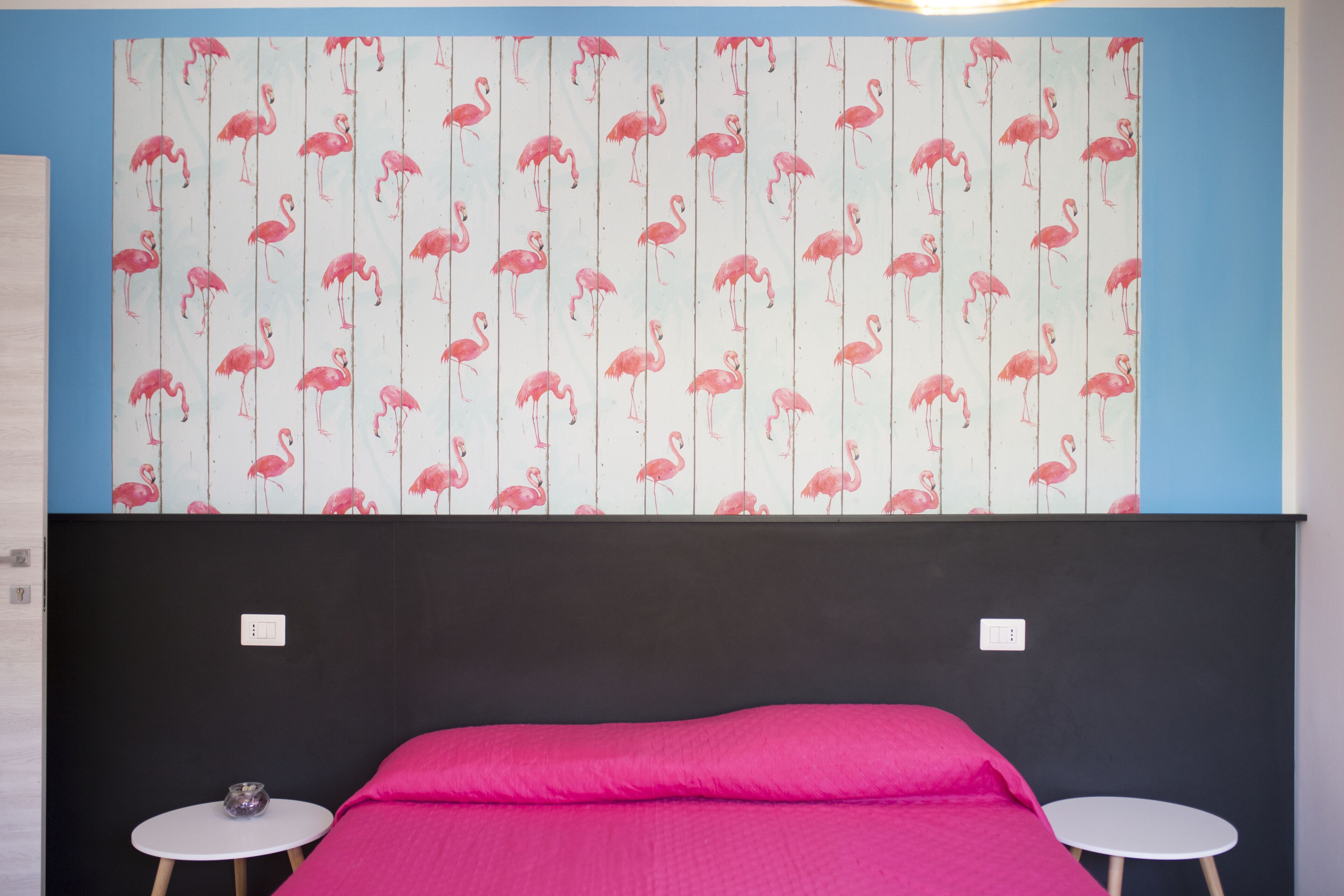 Quarto com uma cama, duas mesinhas redondas e uma parede com padrão de flamingos.